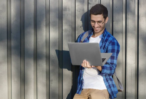 Imagen horizontal al aire libre del joven guapo europeo de pie con la mochila presionada a la cerca de madera gris con el ordenador portátil en las manos, navegar y mirar a través de las páginas web con sonrisa alegre en el día de verano —  Fotos de Stock