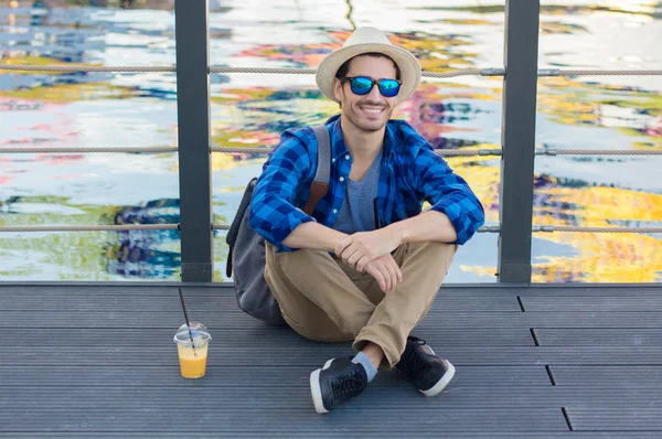 Outdoor portrait of handsome Caucasian guy having rest on wooden surface in park overlooking big beautiful water pond wearing sun hat and sunglasses while looking around and enjoying leisure time — Stock Photo, Image