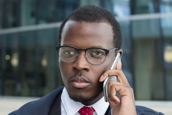 Horizontal headshot of young African businessman pictured in urban environment, dressed in formal suit with tie, wearing eyeglasses and discussing issues on phone with serious face expression