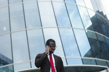 Çalışma konuları tartışmaya yüzünde mutlu gülümseme ile telefonda konuşurken resmi elbise genç yakışıklı Afrikalı-Amerikalı işadamı yüksek şehir bina önünde resim yatay görüntü giymiş