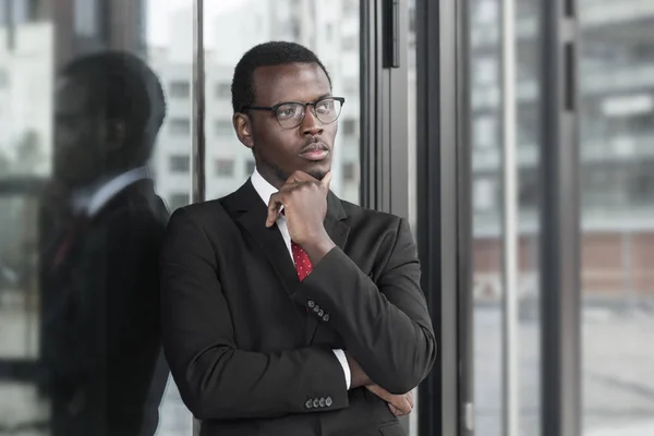 フォーマルな服の彼のオフィスで立って、窓から見て、事業の推進のための新しい戦略を考えて場合、あごを傷に身を包んだ若いアフリカ系アメリカ人男性の画像の水平方向 — ストック写真