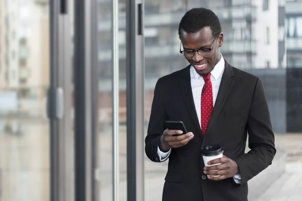 Indoor portrait of busy African chief executive officer pictured in afternoon, holding smartphone in hands and coffee cup, looking attentively at screen with satisfied happy smile while checking web