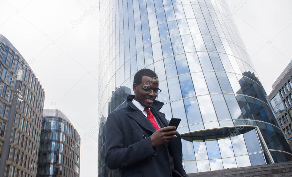 Urban portrait of African male standing with high glass building in background dressed in formal clothes, looking attentively at screen of phone showing friendly smile and interest in business news