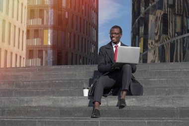 Yatay görüntü Caddesi'ne olumlu gülümseme ile görünen resmi kıyafet ve şehir merkezinde, merdivenlerde otururken dizüstü kullanarak gözlük yakışıklı esmer adam duygu rahat ve mutlu