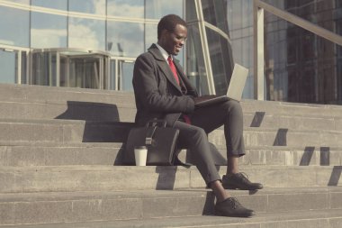 Afrikalı girişimci koyu renkli şehir merkezinde merdivenlerde molası sırasında oturan yatay görüntü giyinmiş resmi kıyafetler, kahve içer ve diz üstü konsantre bakışla işi