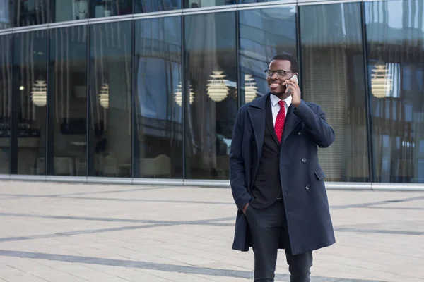 Horizontal photo of young dark-skinned African businessman standing in street in city center with glass window walls behind, answering phone call from his office, helping colleagues with work