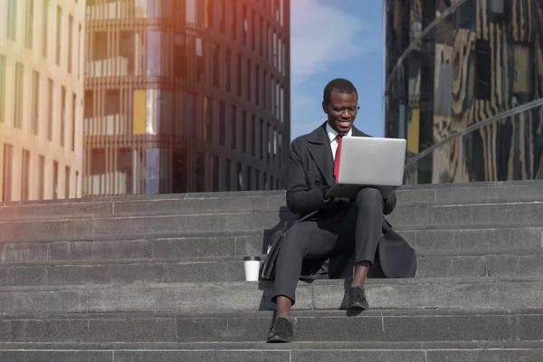 Horizontal closeup of young African American businessman sitting outdoors on stairs with laptop open feeling joyful and comfortable while looking at content on open laptop, smiling positively