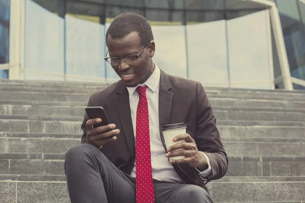 Foto urbana de un joven hombre de negocios afroamericano guapo que pasa tiempo libre en las escaleras en la calle bebiendo café para llevar y revisando mensajes o comunicándose a través de un teléfono inteligente con sonrisa —  Fotos de Stock