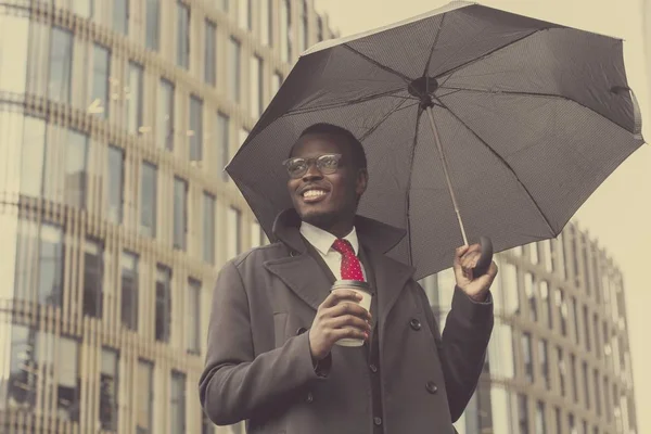 Urban portrait of handsome African American businessman standing in city center in cloudy weather under black umbrella dressed in formal clothes, wearing glasses and smiling happily with coffee