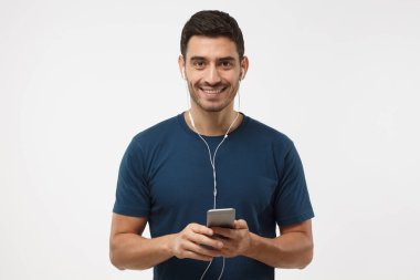 Gri arka plan üzerinde izole müzik dinlerken kameraya, seyir kulaklıklar smartphone, holding gülümseyen yakışıklı erkek portresi