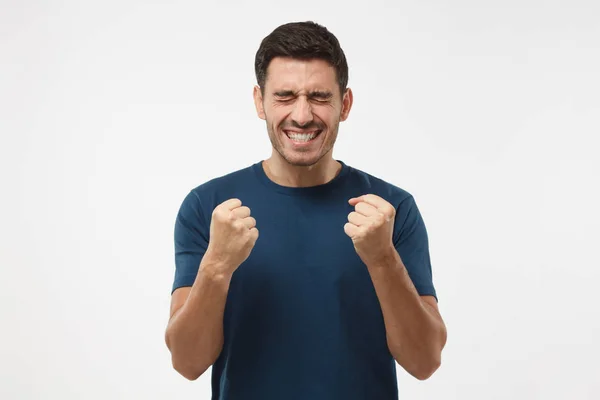 Primer plano del hombre europeo emocional aislado sobre fondo gris que muestra dientes blancos mientras grita con alegría y expresión victoriosa, tomados de la mano en gesto de ganador, mirando extremadamente feliz — Foto de Stock