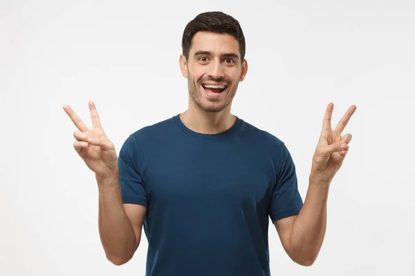 Indoor portrait of young male isolated on gray background in casual clothes with optimistic smile, showing victory sign with both hands, looking friendly and willing to welcome and communicate — Stok fotoğraf