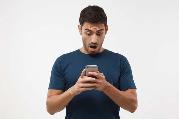 Nahaufnahme Porträt eines hübschen jungen Mannes in blauem T-Shirt, der sein Smartphone in der Hand hält, verängstigt und verwirrt schaut, als er SMS oder E-Mail liest — Stockfoto