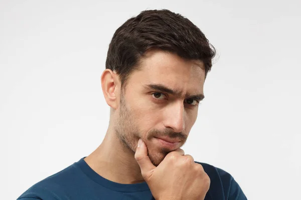 Close up portrait of young handsome male in blue t-shirt touching his chin with hand thoughtfully looking at camera. Doubt concept — Stock Photo, Image