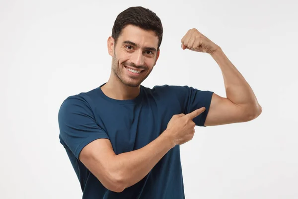 Joven hombre deportivo fuerte en camiseta azul, muestra bíceps después del entrenamiento en el gimnasio, aislado sobre fondo gris — Foto de Stock