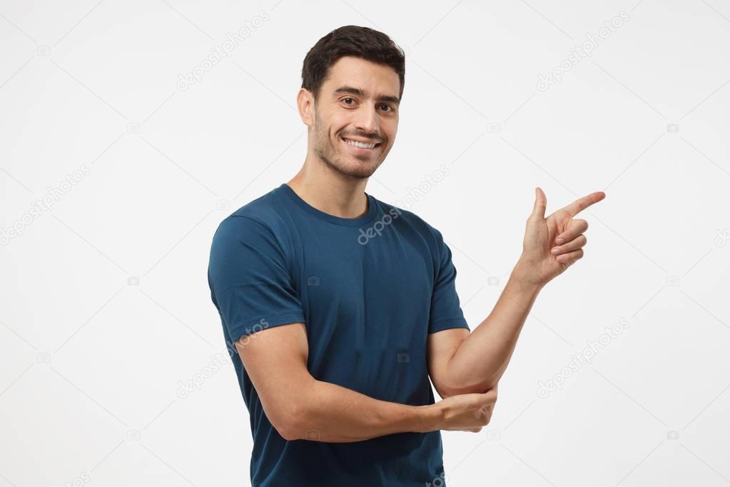 Attractive young man in blue t-shirt pointing right with his finger isolated on gray background