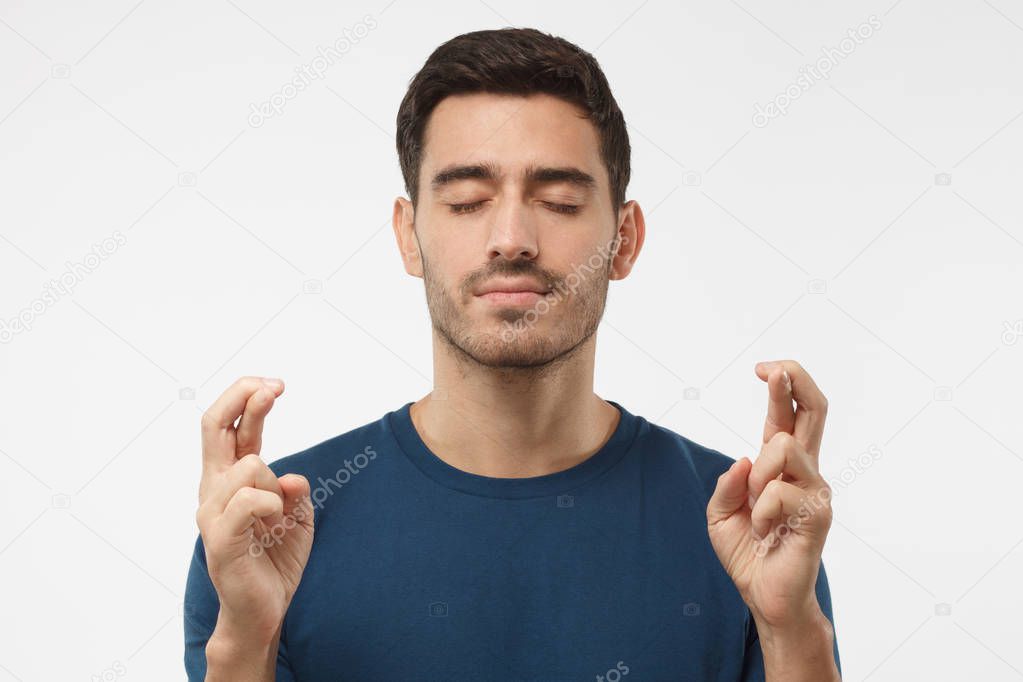 Indoor photo of young man isolated on grey background, looking deeply concentrated with eyes closed and fingers of both hands crossed, wishing luck, praying