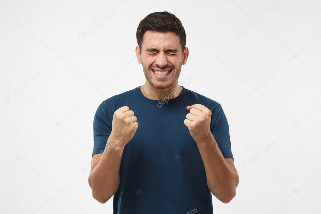 Closeup of emotional European man isolated on gray background showing white teeth while screaming with joy and victorious expression, holding hands in gesture of winner, looking extremely happy