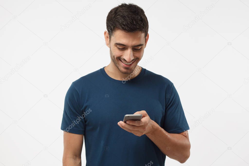 Indoor shot of good-looking young male isolated on gray background looking at smartphone, smiling openly while holding smartphone in one hand 