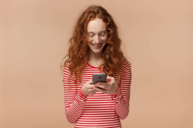 Çizgili tişört cep telefonu ekranına dikkatle bakarak, web sayfalarını tarama ve güzel gülüyor arkadaşlarınızla sohbet ederken gülümseyen çekici genç Kızıl saçlı erkek