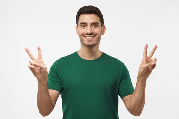 Joven hombre guapo aislado sobre fondo gris en camiseta verde con sonrisa optimista, mostrando signo de victoria con ambas manos, buscando amigable — Foto de Stock