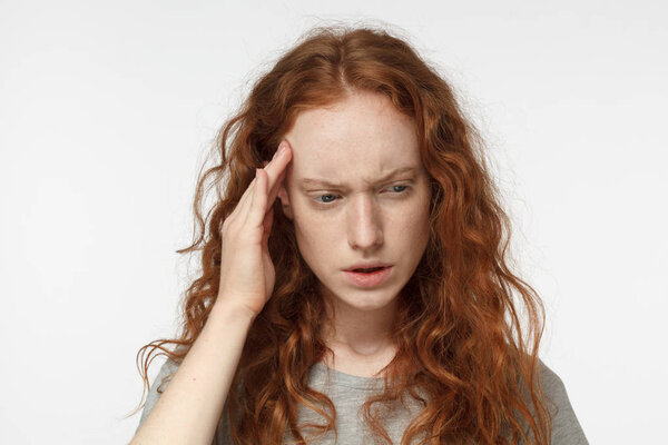 Indoor portrait of young beautiful redhead European female isolated on white background frowning her eyebrows and pressing fingers to her temple as if experiencing migraine, feeling sick and stressed