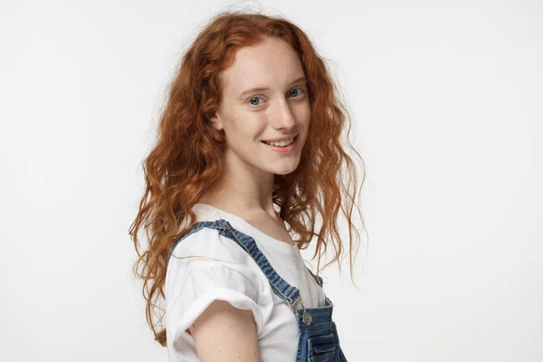 Closeup photo of young teenage girl with red loose hair pictured isolated on white background dressed in white T-shirt and denim jumpsuit, turned to camera with her shoulder, smiling cheerfully — Stock Photo, Image