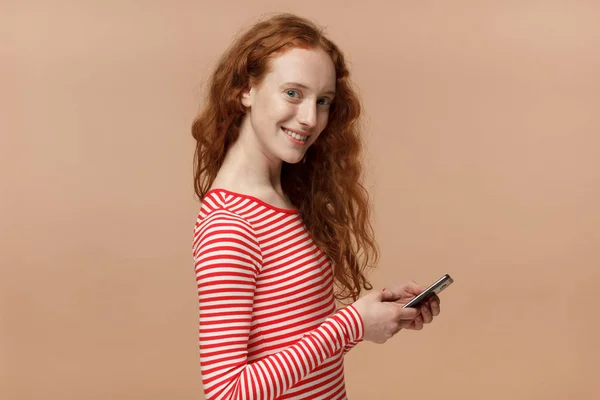 Menina ruiva muito encaracolado em t-shirt listrada olhando e sorrindo para a câmera com expressão alegre ao enviar mensagens de texto para seus amigos, usando telefone celular, isolado no fundo pêssego — Fotografia de Stock