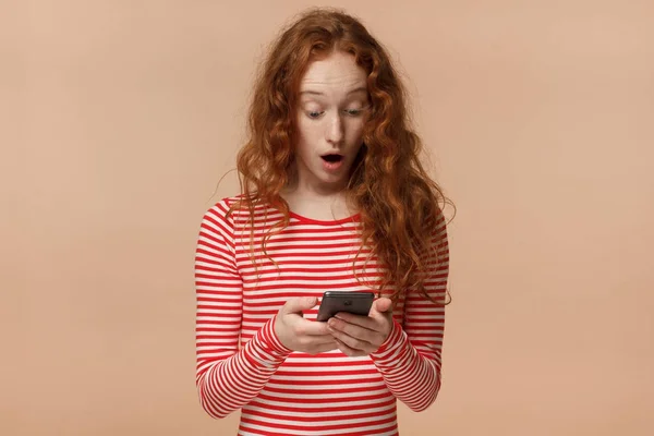 Retrato de atraente jovem ruiva fêmea, segurando smartphone, olhando assustado e confuso leitura mensagem de texto ou e-mail — Fotografia de Stock