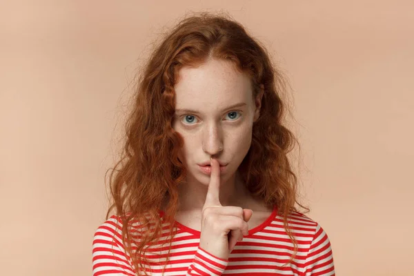 Primo piano colpo di attraente giovane rossa con gesto shhh, chiedendo silenzio o di essere tranquillo, isolato su sfondo beige — Foto Stock