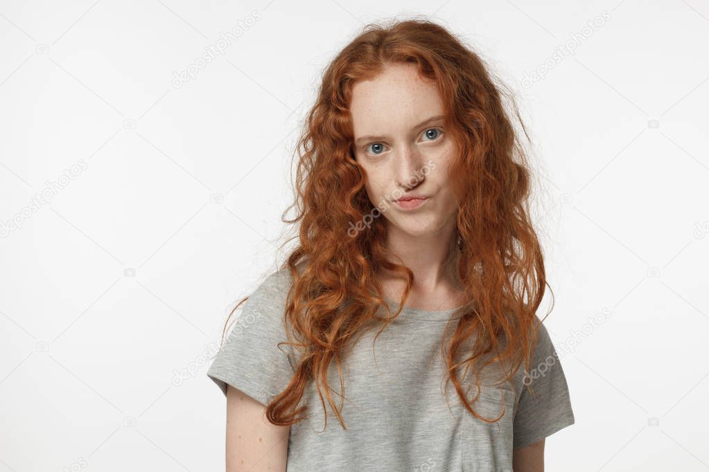 Horizontal portrait of young attractive European woman with red hair isolated on white background, wearing grey T-shirt, her lips twisted in sign of disagreement, doubt, dissatisfaction and mistrust