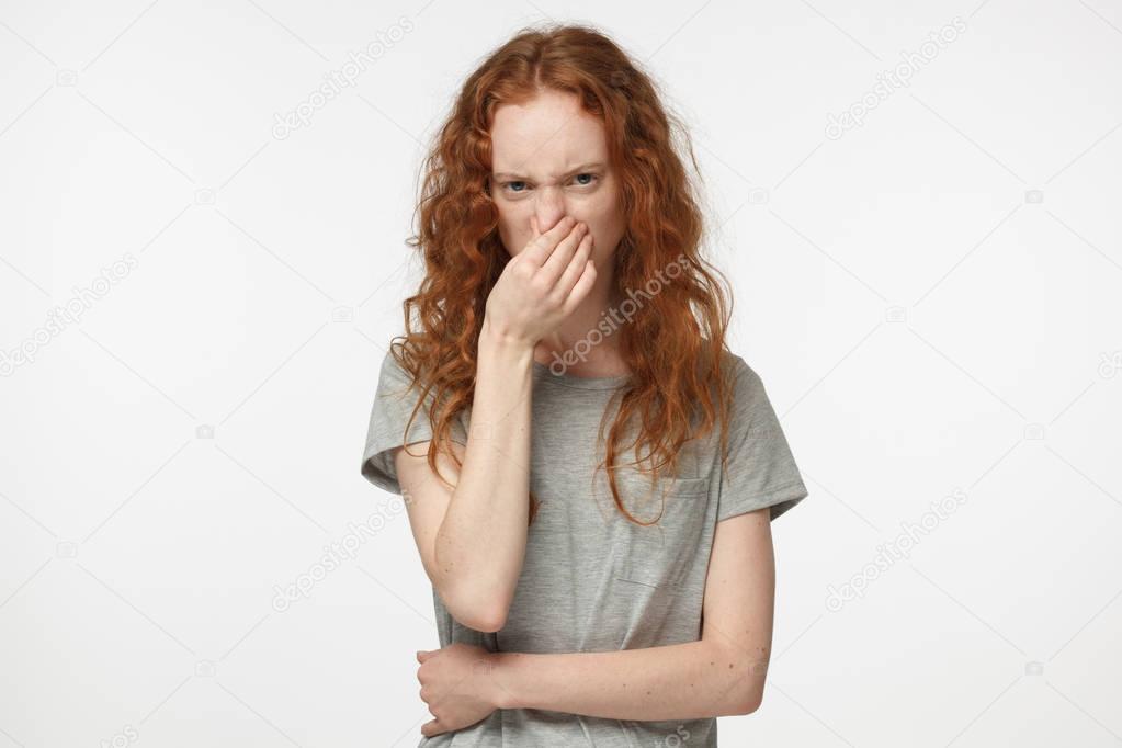 Closeup headshot of young attractive European woman with red hair isolated on white background holding her nose with wry face showing deep dissatisfaction and disgust with unpleasant smell or facts