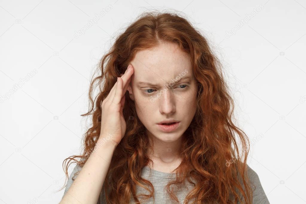 Indoor portrait of young beautiful redhead European female isolated on white background frowning her eyebrows and pressing fingers to her temple as if experiencing migraine, feeling sick and stressed