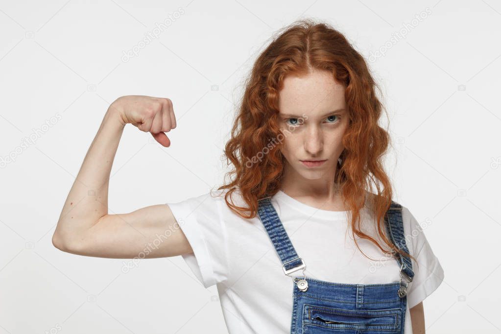 Indoor portrait of young beautiful redhead European girl isolated on white background with stubborn and fierce look in eyes showing biceps muscle having lifted arm with clenched fist to prove strength