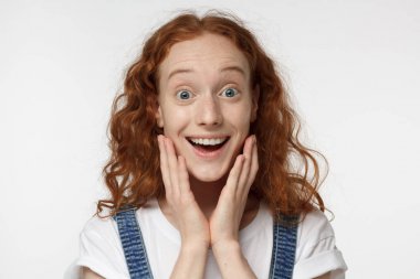 Horizontal headshot of young beautiful redhead European female isolated on white background screaming happily in great astonishment, her eyes and mouth open eyed, palms pressed to her cheeks clipart