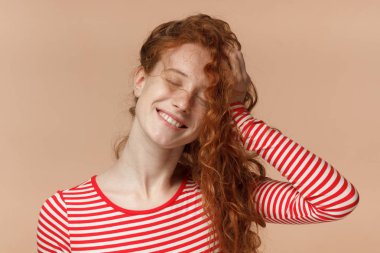Yatay resim üzerinde şeftali arka plan izole çekici Kızıl saçlı Bayan dalgalı saçları ile karışıklık yapmak, duygu rahat ve mutlu çizgili tişört giymiş genç ile gülümseyen gözleri kapalı