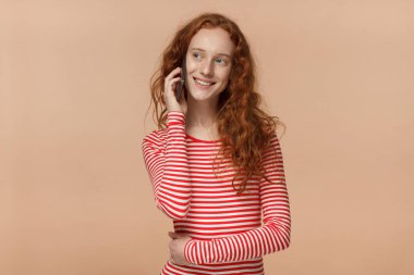 Yatay resim smartphone kulak, yanındaki tutarak rahat kıyafetler giymiş şeftali arka plan üzerinde izole genç çekici Kızıl saçlı Bayan sidewards gülümseme ile arkadaşıyla konuşurken arıyorum