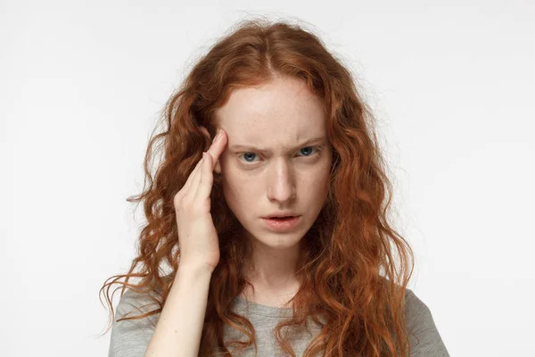 Lähikuva nuoresta viehättävästä eurooppalaisesta naisesta, jolla on valkoisella taustalla punaiset hiukset ja joka koskettaa ohimoaan sormilla kuin yrittäessään muistaa jotain tärkeää tai keskittyä ongelmaan — kuvapankkivalokuva