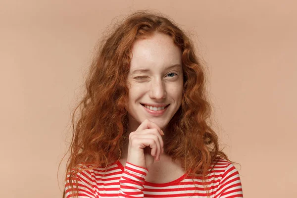 Studio headshot of good-looking redhead teen girl isolated on peach background looking enterprising and enthusiastic, winking friendly as if inviting to adventure or recommending good benefit