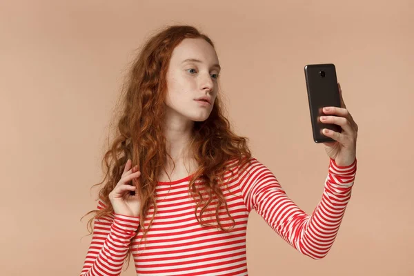 Estúdio headshot de jovem ruiva atraente senhora europeia isolada em fundo pêssego usando câmera frontal de celular para tirar foto selfie, posando como modelo, a fim de compartilhar imagem na web — Fotografia de Stock