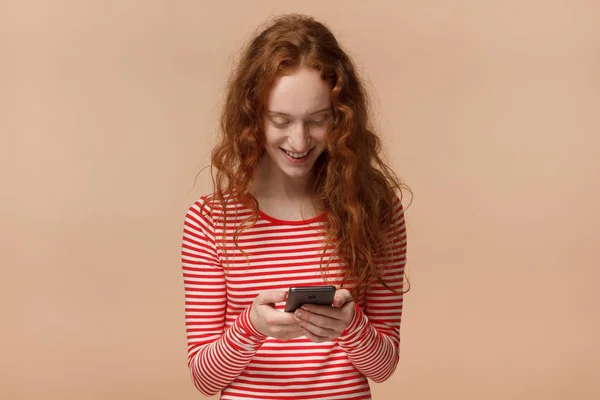 年轻漂亮的欧洲妇女与红松散卷曲的头发在桃背景下的室内图片手持电话, 在浏览或玩游戏时, 要用心地看屏幕 — 图库照片