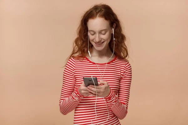水平照片的年轻漂亮红发欧洲女人孤立在桃背景手持手机, 打字信息或选择音轨, 而听音乐通过耳机 — 图库照片