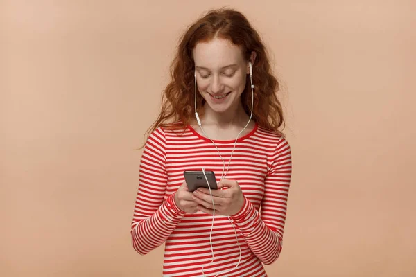 Foto horizontal de jovem ruiva bonita mulher europeia isolada no fundo pêssego segurando celular em mãos, digitando mensagens ou escolhendo faixas de áudio enquanto ouve música através de fones de ouvido — Fotografia de Stock