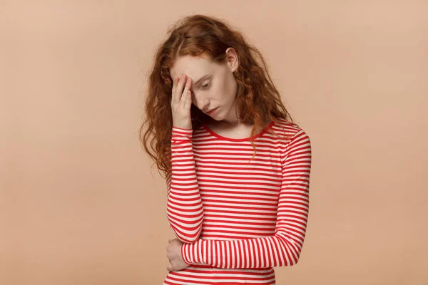 Studio photo of young attractive European lady with red curly hair isolated on peach background pressing palm to forehead looking worried, bored and exhausted with hard mind work or personal trouble
