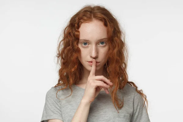 Primo piano colpo di attraente rossa ragazza riccia con gesto shhh, chiedendo silenzio o di essere tranquillo, isolato su sfondo grigio — Foto Stock