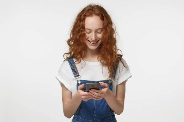 Vnitřní záběr hezká mladá zrzka žena izolována na šedém pozadí pohledu na smartphone, usmívající se otevřeně přitom v obou rukou držel smartphone — Stock fotografie