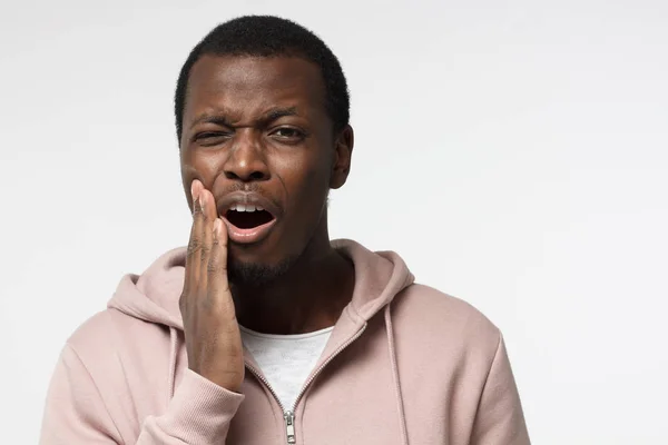 Jonge zwarte man geïsoleerd op grijze achtergrond wihh mond open, het aanraken van zijn gezicht met expressie van verschrikkelijk lijden gezondheidsprobleem en pijnlijke tand — Stockfoto