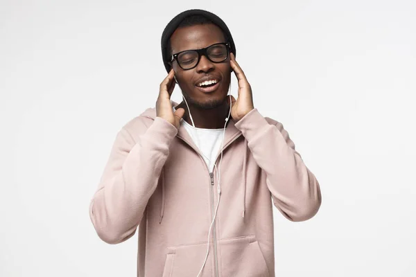 Porträt eines glücklichen jungen schwarzen Mannes in rosa Kapuzenpulli, der mit beiden Händen Kopfhörer hält; Musik mit geschlossenen Augen hört; isoliert auf grauem Hintergrund — Stockfoto
