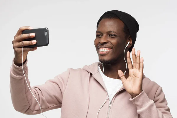 Zeg hallo! Portret van de jonge knappe Afrikaanse man die tegen de grijze achtergrond via smartphone op selfie foto's en lachend — Stockfoto