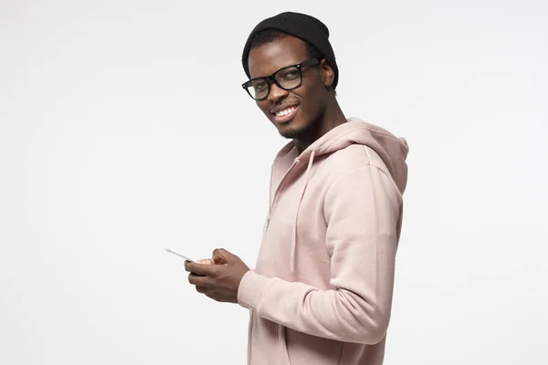 Schwarzer Kerl, der mit fröhlichem Gesichtsausdruck in die Kamera schaut und lächelt, während er seinen Freunden SMS schreibt, mit Handy, isoliert auf grauem Hintergrund — Stockfoto